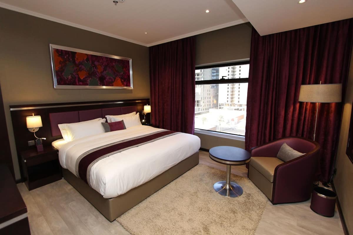A hotel room with a large bed, a small round table, a chair, a nightstand with a lamp, and a window with curtains.