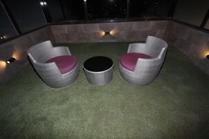 Un área de estar en la azotea con dos sillones de mimbre y una mesa redonda, rodeada de un piso de césped artificial y paredes de piedra.