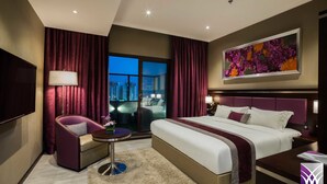 A modern hotel room with a large bed, a seating area with a chair and table, a flat-screen TV, and a city view through the window. - Normas Hotel (Al Khobar)