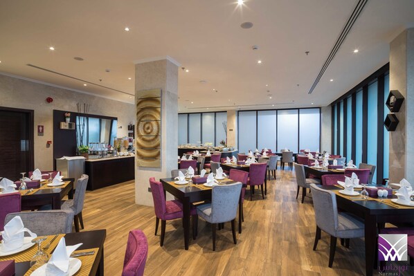 Un restaurante espacioso con varias mesas y sillas, una zona de bar central y grandes ventanales que dejan entrar luz natural.