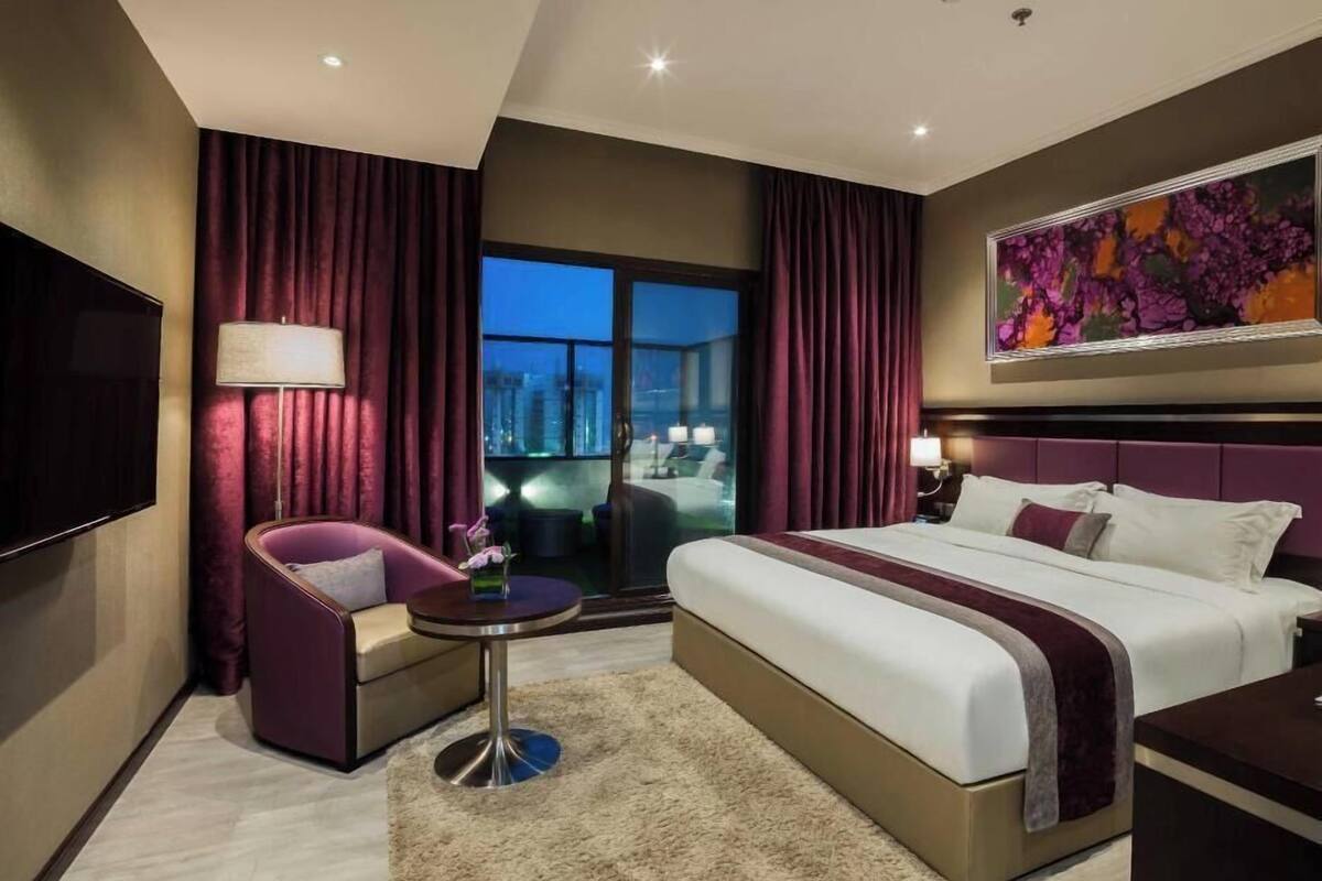 A hotel room with a bed, a seating area with a table, a TV, and a large window with curtains.