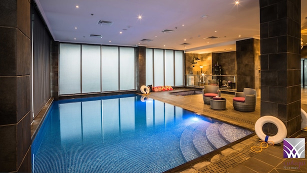 A modern indoor swimming pool with clear blue water, surrounded by dark stone walls and a seating area with cushioned chairs. - Normas Hotel (Al Khobar)