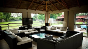 Een ruime woonkamer met rieten dak, zachte zitmogelijkheden, een flatscreen televisie en uitzicht op een zwembad en groen.