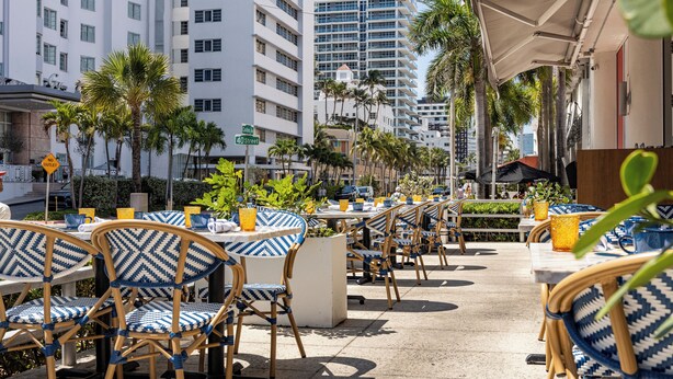 Outdoor seating with patterned chairs and tables, palm trees, and modern buildings in the background. - Hotel Continental Miami Beach, Tapestry Collection by Hilton (Miami Beach)
