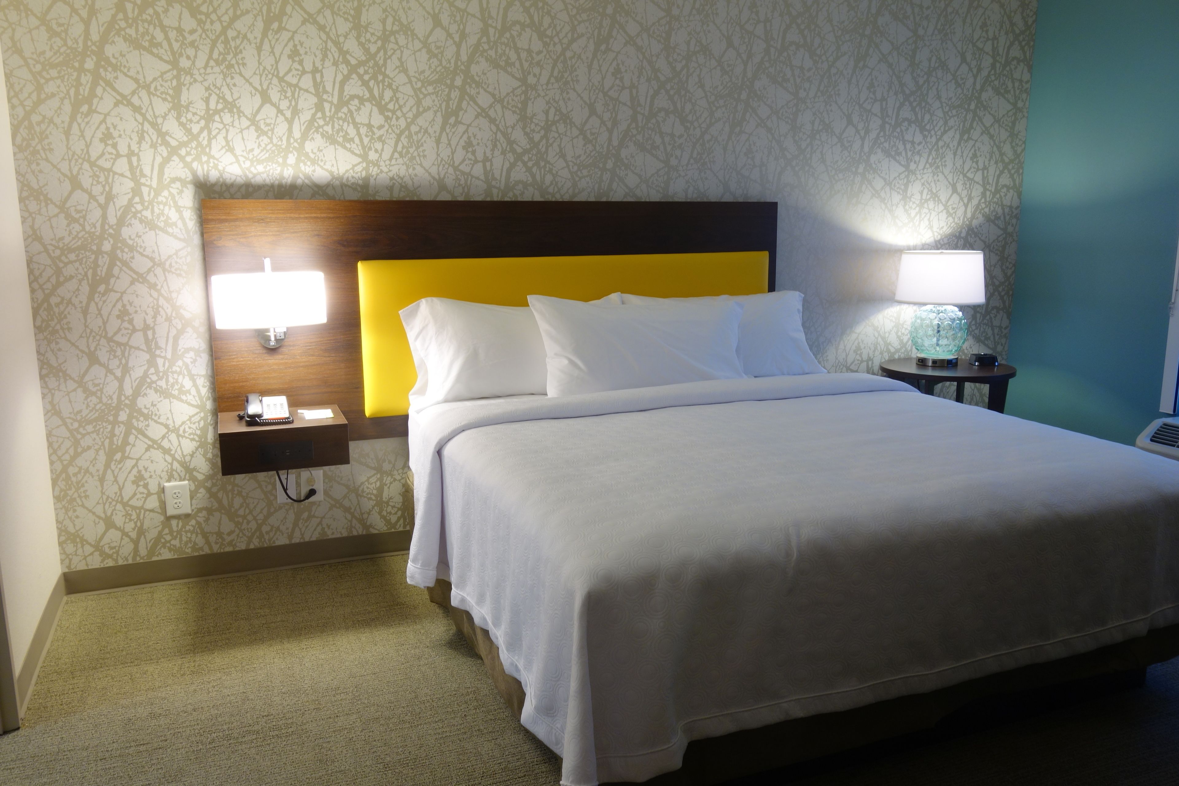 Habitación de hotel con una cama bien hecha, una mesita de noche con lámpara y un teléfono en la pared.