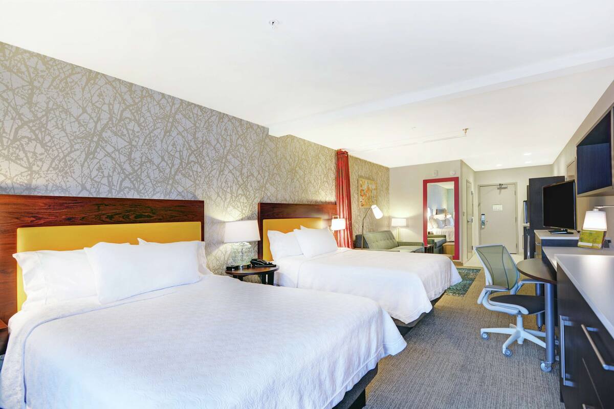 A hotel room with two beds, a desk, a chair, and a TV.