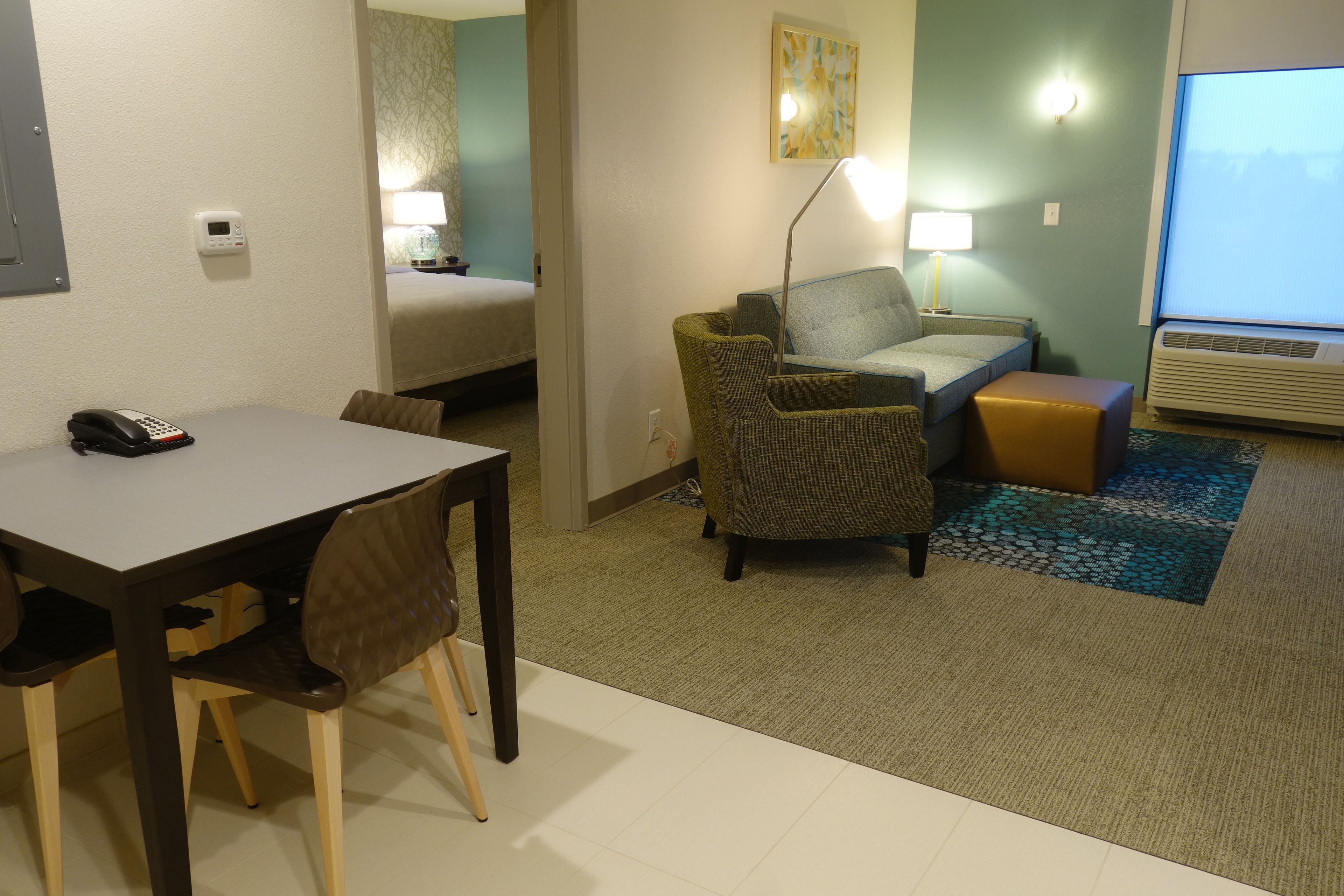 Et hotelværelse med et spisebord, stole, en sofa, en lille skammel, en lampe, et billede og et vindue med udsigt.