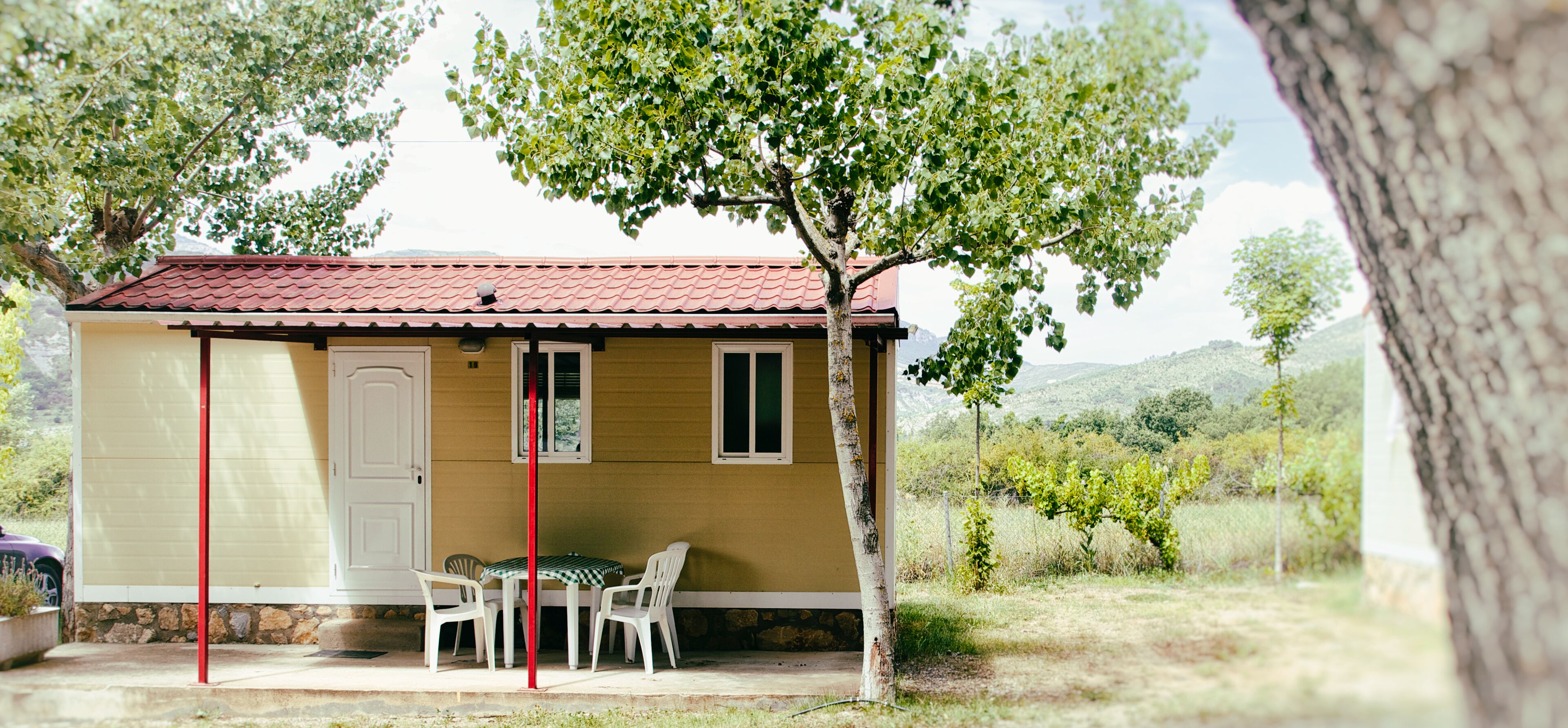Une petite maison jaune avec un toit rouge, une porte blanche et une petite table avec deux chaises à l’extérieur.