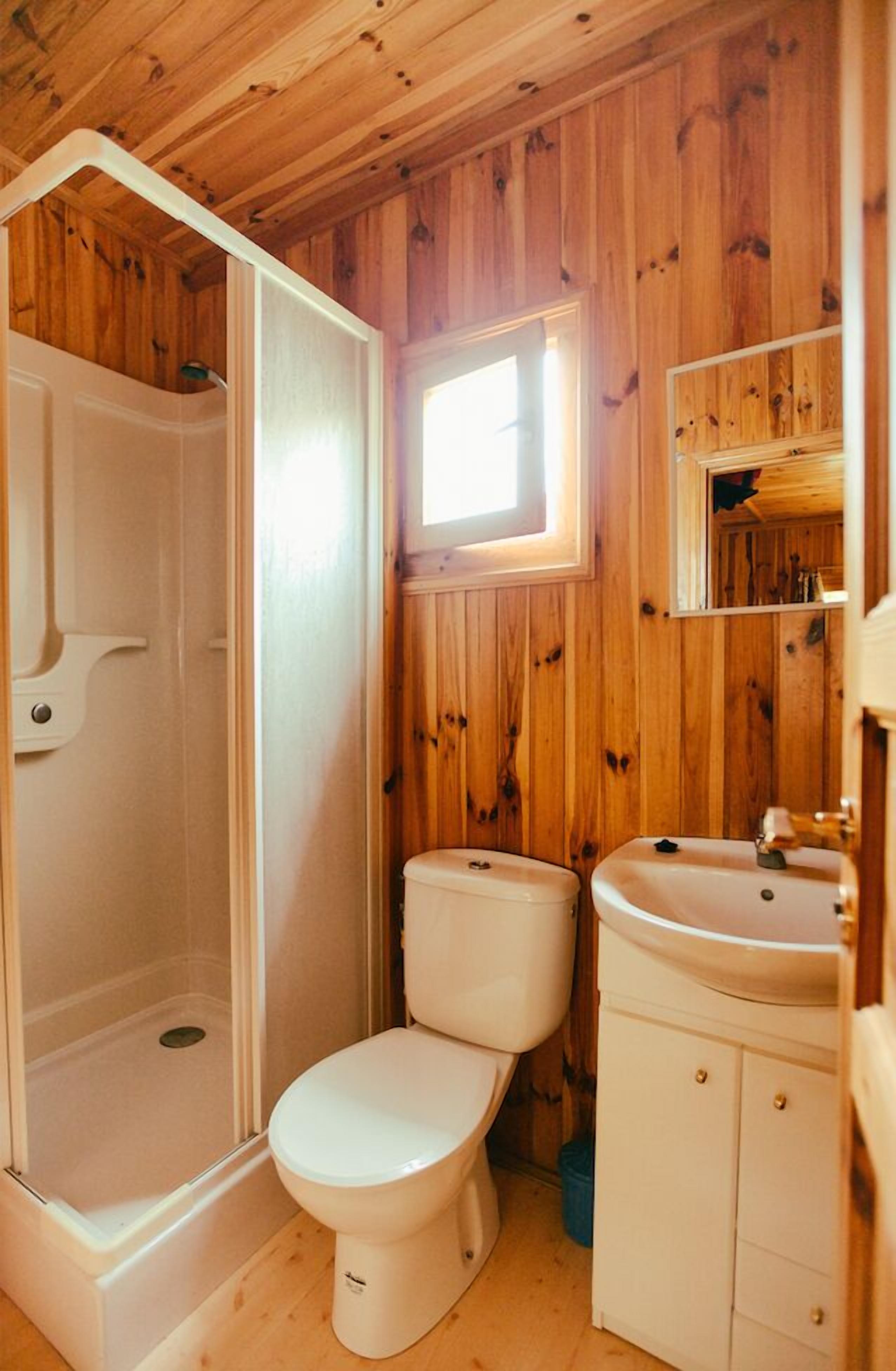 A compact bathroom with wooden walls, a white toilet, a sink, and a shower enclosure.
