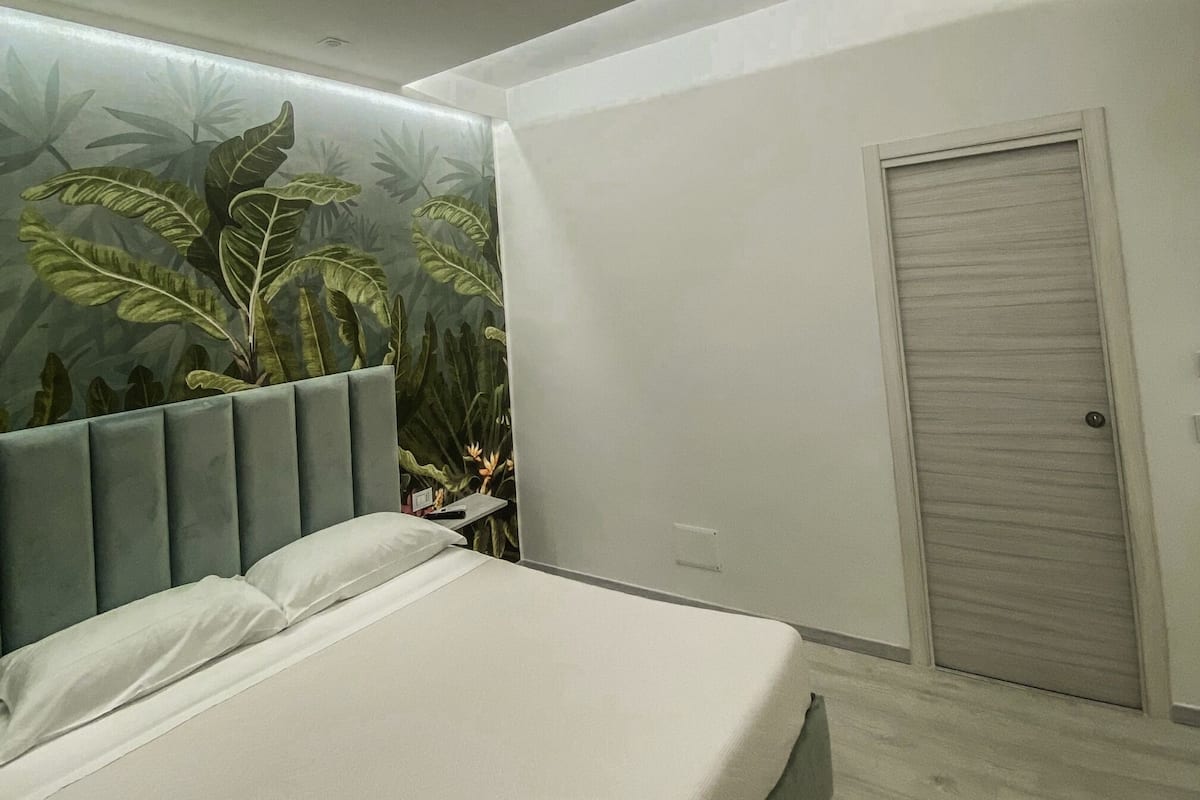 A modern bedroom with a bed, a wall with a tropical plant mural, a door, and a ceiling light.