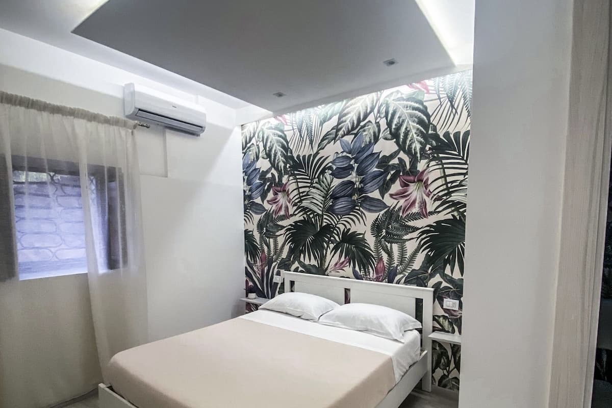 A bedroom with a bed, a window with curtains, an air conditioning unit, and a wall with a tropical leaf pattern.