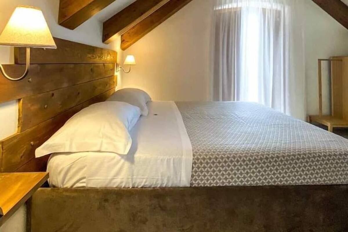 A bedroom with a sloped ceiling, wooden beams, a bed with white bedding, and a bedside lamp.