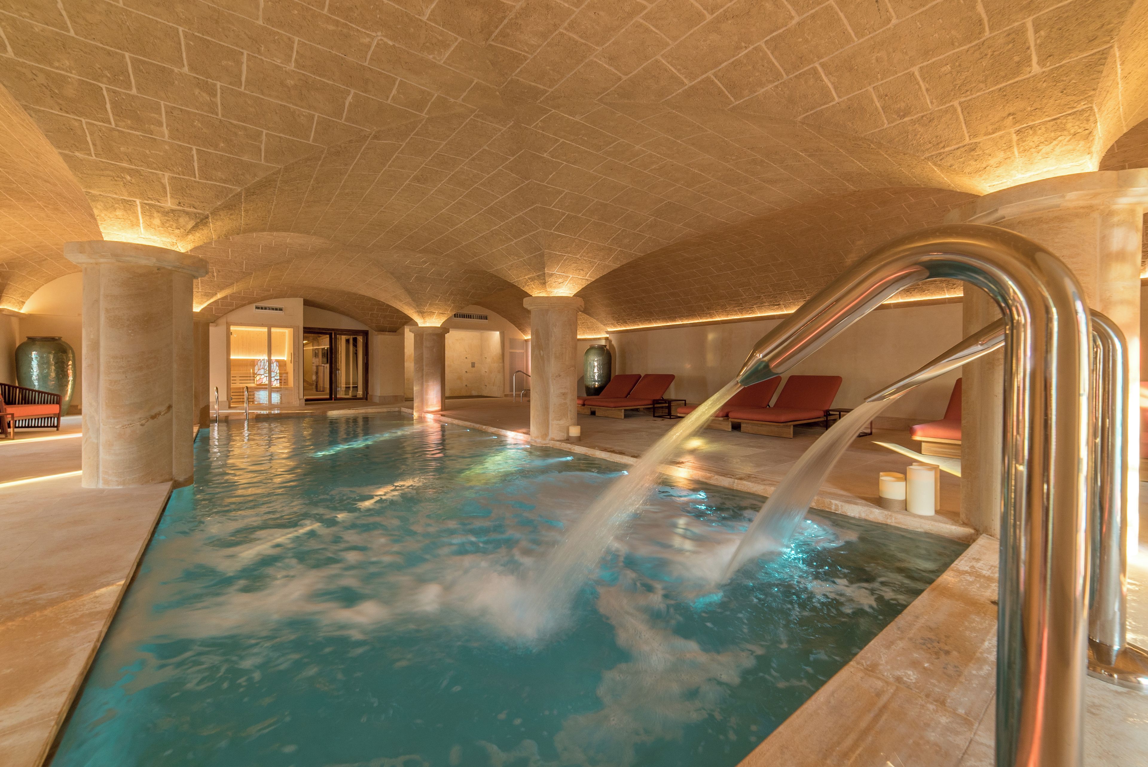Una piscina interior con fuente de agua, columnas de piedra y sillas de descanso rojas.