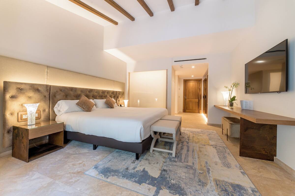 A modern bedroom with a large bed, bedside tables, a bench, a TV, and a wooden ceiling beam.