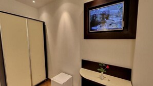 A hotel room with a television mounted on the wall, a small white cabinet, and a bedside table with a vase of flowers. - Ikram el Dhayf (Algiers)