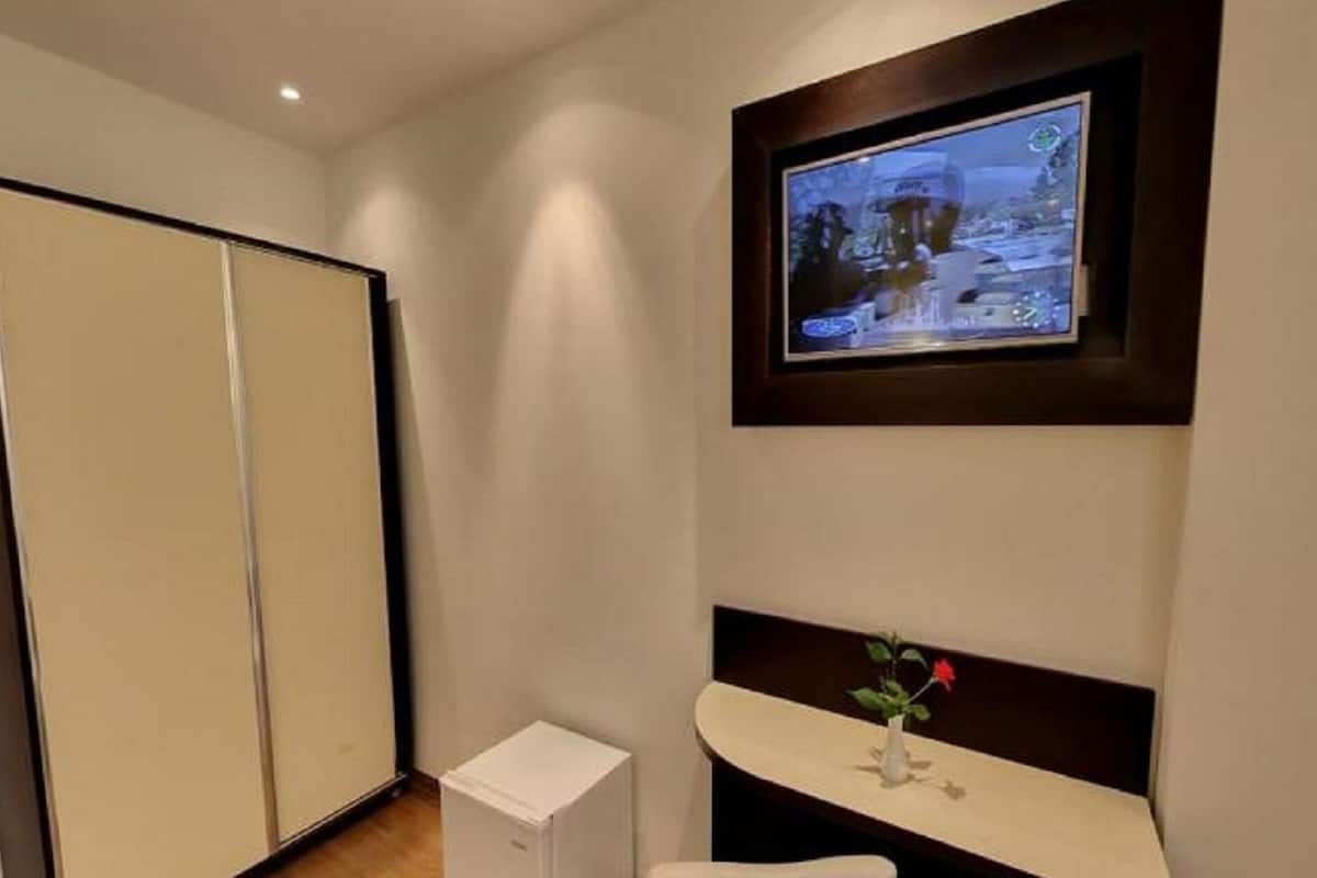 A hotel room with a television mounted on the wall, a small white cabinet, and a vase with flowers on a shelf.