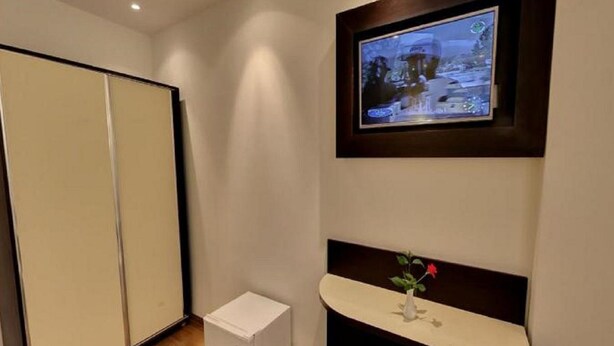 Habitación de hotel con televisor en la pared, un mueble blanco pequeño y una mesita de noche con un florero.