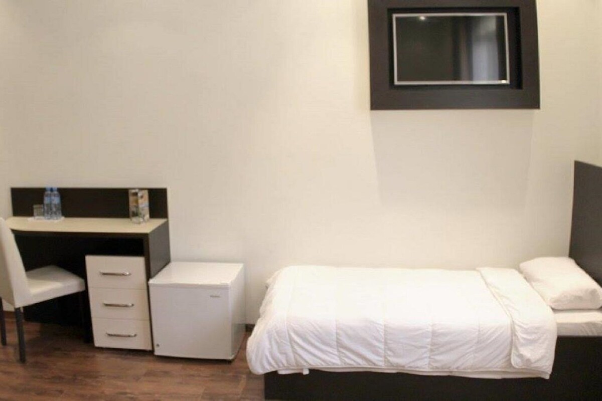 A compact hotel room with a single bed, a desk with a chair, and a small bedside table.