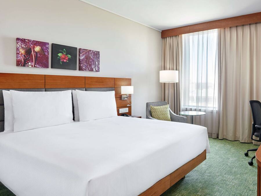 Habitación de hotel con una cama grande, un escritorio con computadora, una silla, una mesita y una ventana con cortinas.