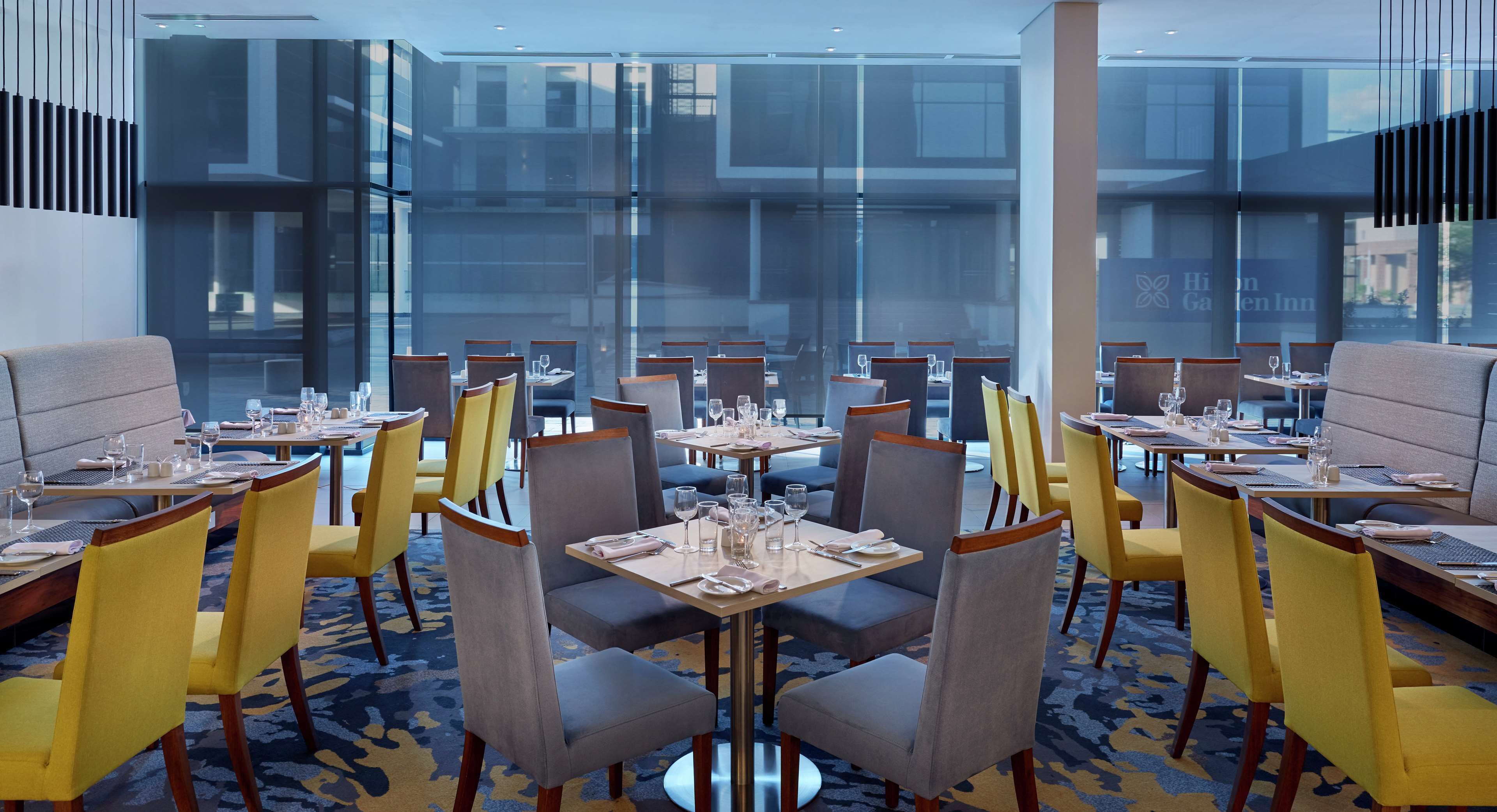 Un restaurante moderno con mesas dispuestas para comer, con una combinación de sillas amarillas y grises, y grandes ventanales que ofrecen vistas del paisaje urbano.