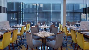 Un restaurante moderno con mesas dispuestas para comer, con una combinación de sillas amarillas y grises, y grandes ventanales que ofrecen vistas del paisaje urbano.