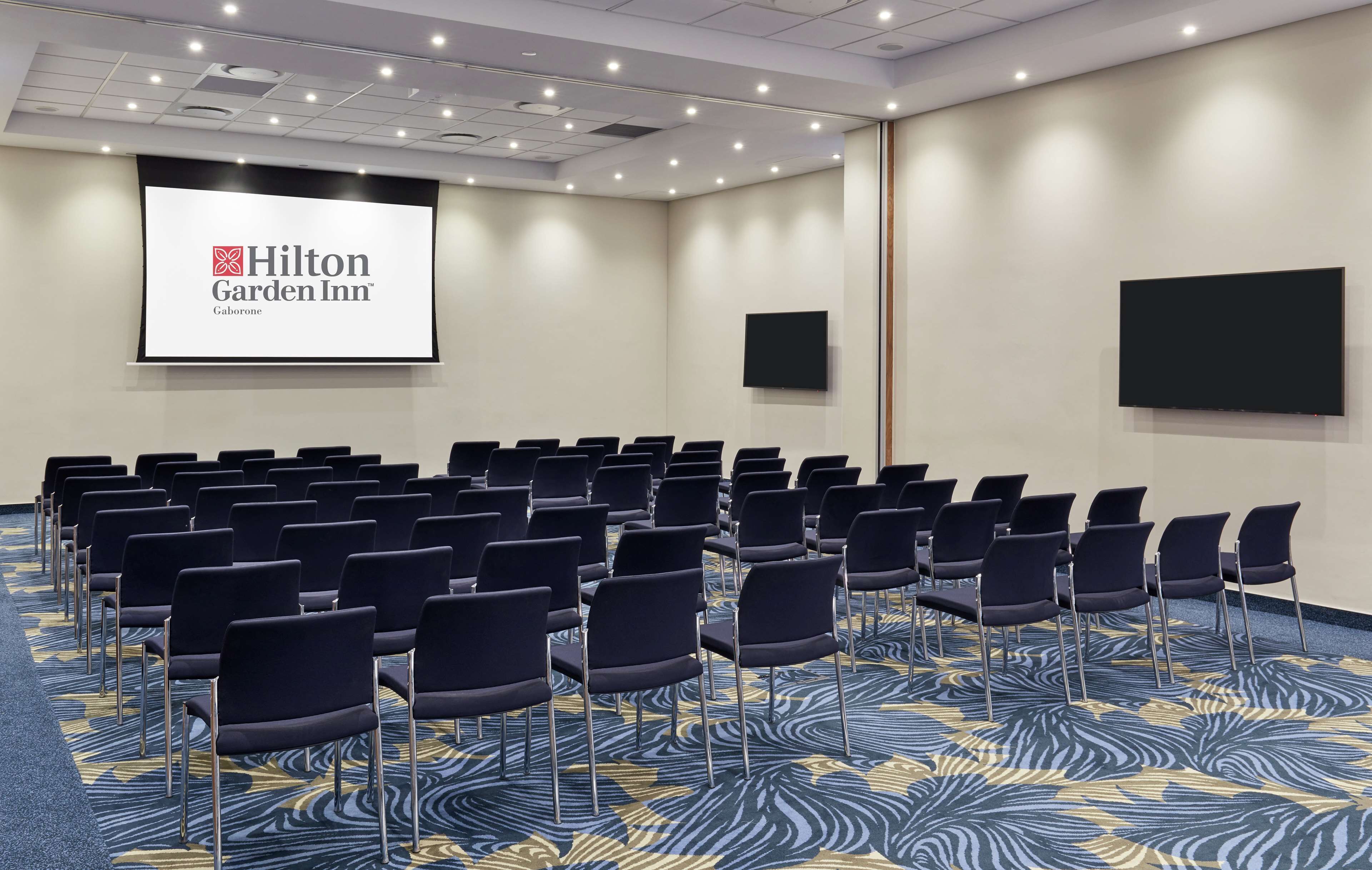 A conference room with rows of chairs, a large screen displaying the Hilton Garden Inn logo, and a carpet with a blue and yellow pattern.