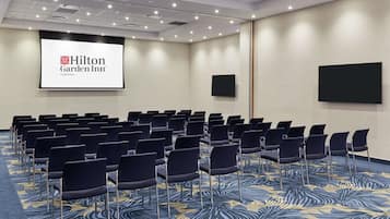 A conference room with rows of chairs, a large screen displaying the Hilton Garden Inn logo, and a carpet with a blue and yellow pattern.
