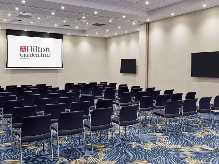 A conference room with rows of chairs, a large screen displaying the Hilton Garden Inn logo, and a carpet with a blue and yellow pattern.