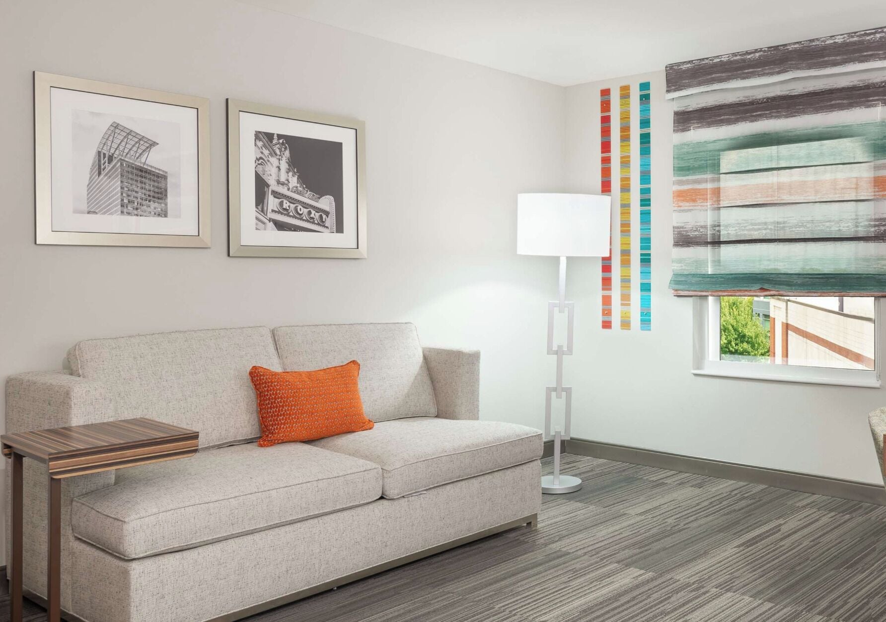 A modern living room with a grey sofa, a small wooden side table, a floor lamp, and two framed pictures on the wall.