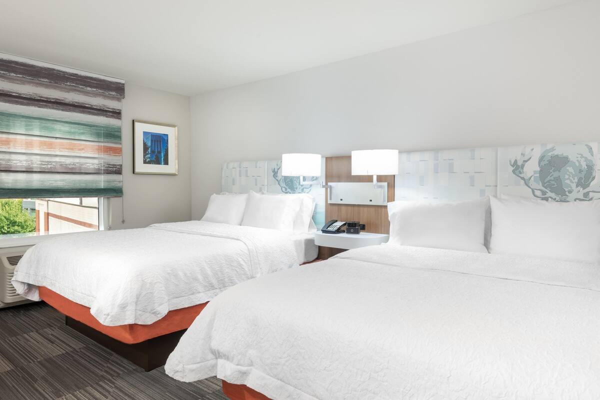 Habitación de hotel con dos camas, un ventanal con persianas, un cuadro en la pared y una mesita de noche con un teléfono y una lámpara.