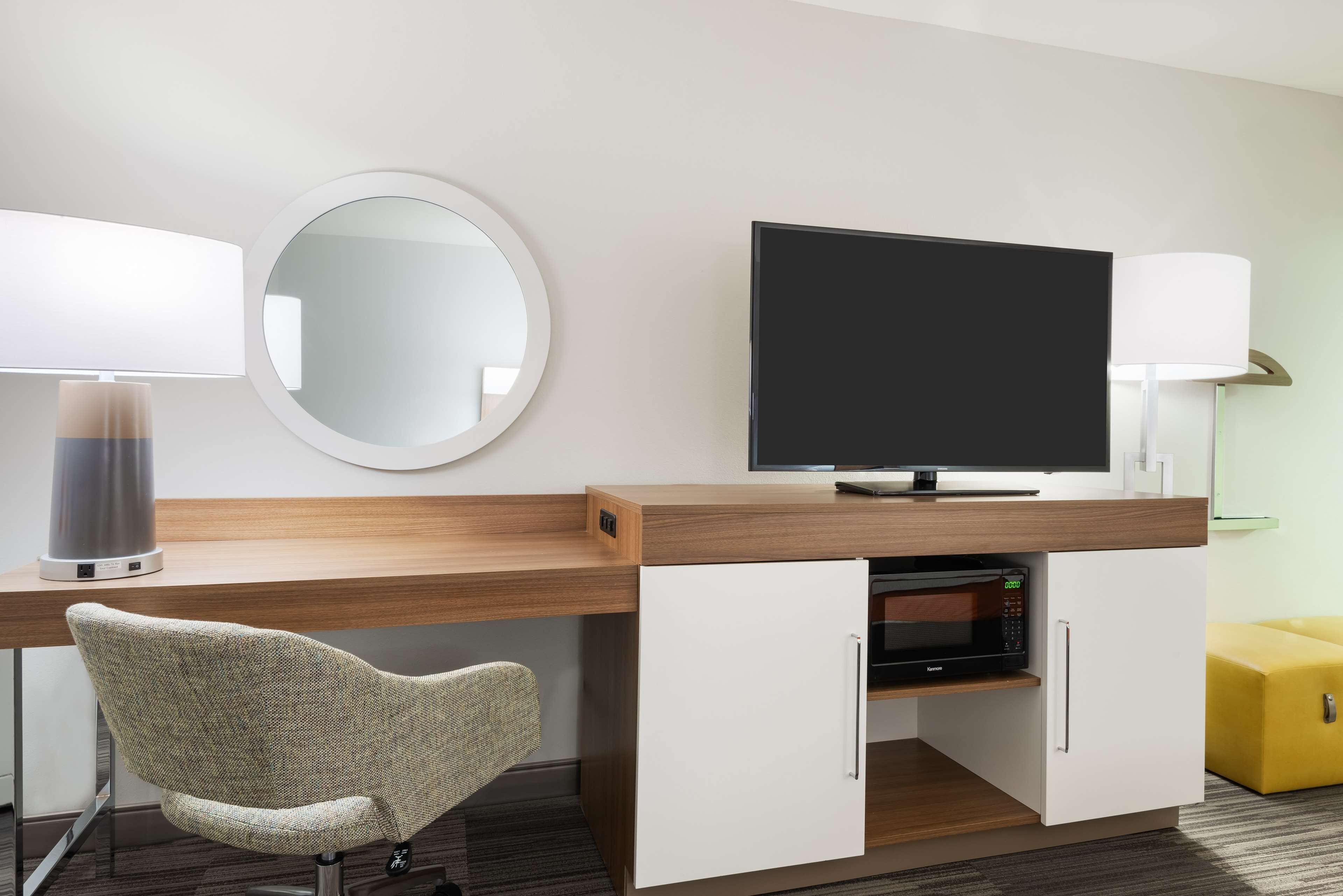 A modern hotel room with a wooden desk, a flat-screen TV, a round mirror, a lamp, a microwave, and a yellow ottoman.