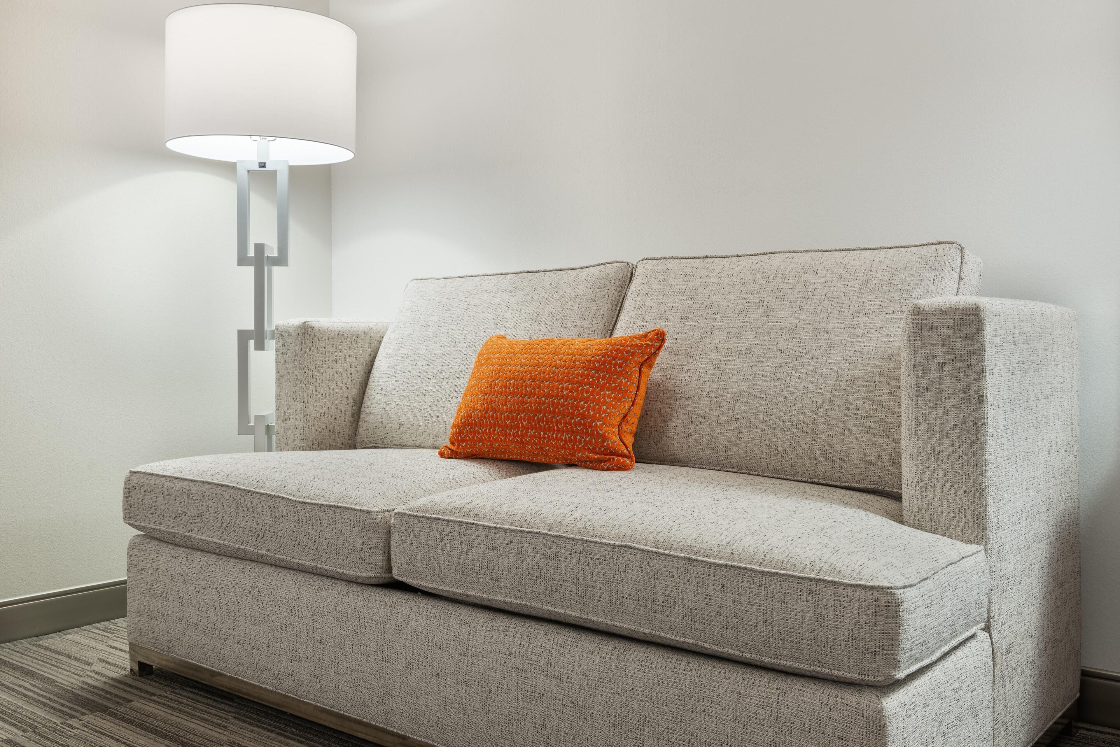 Un canapé gris recouvert d’un tissu texturé, un seul coussin orange et une lampe de sol moderne avec un abat-jour blanc.