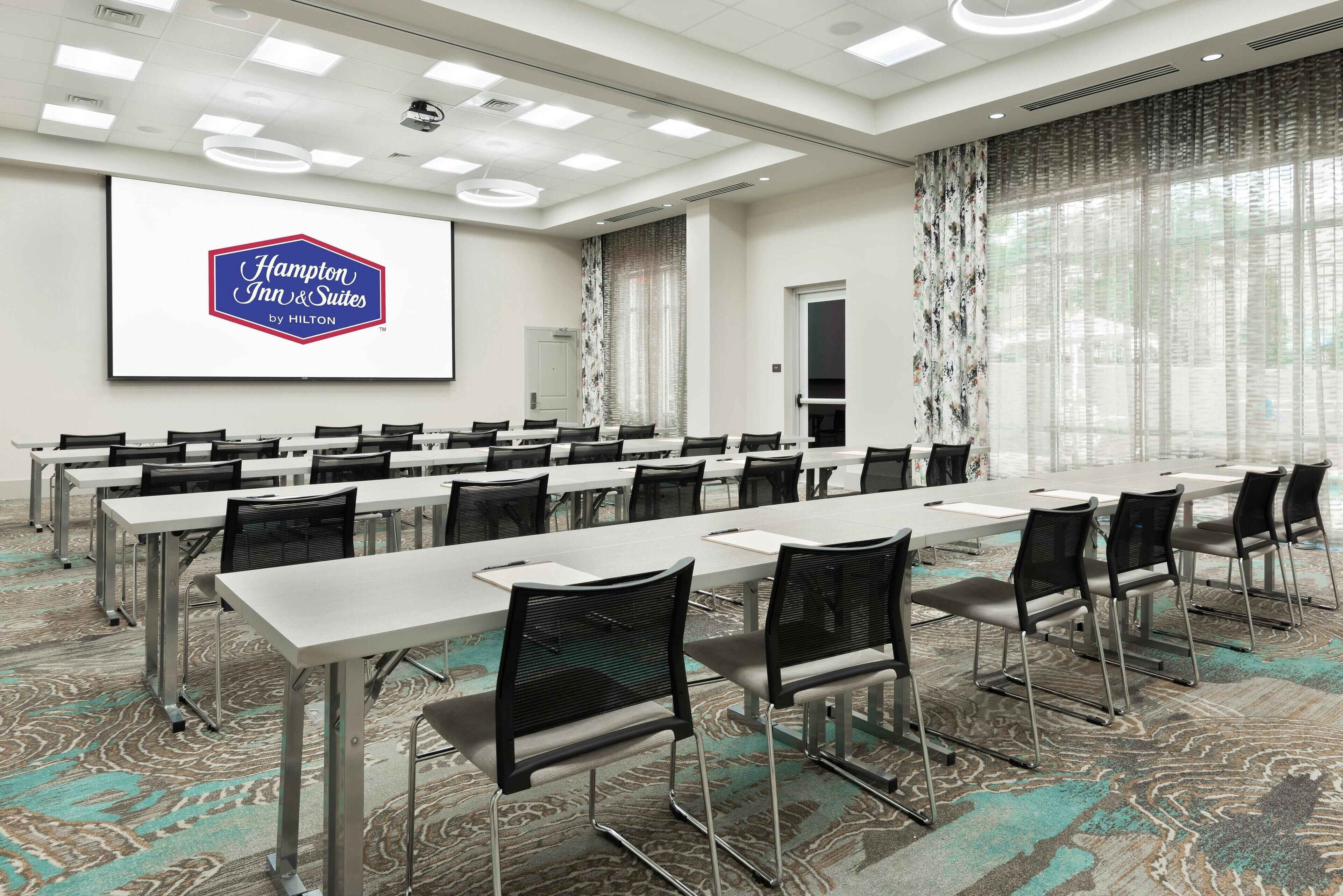 Una sala conferenze con lunghi tavoli e sedie, un grande schermo che mostra il logo Hampton Inn & Suites by Hilton e un tappeto decorato.