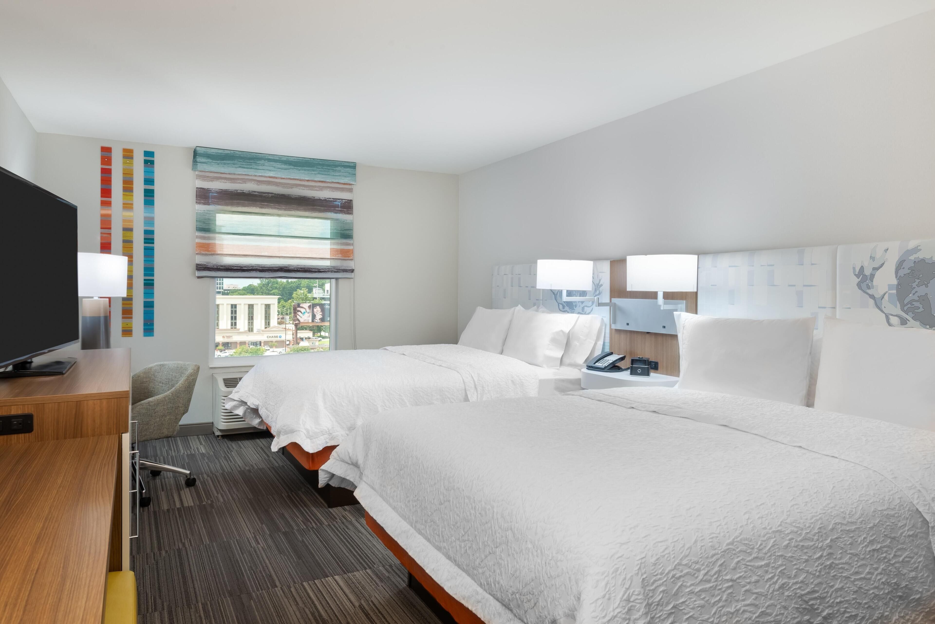 A hotel room with two beds, a desk, a chair, a TV, and a window with a view of buildings.