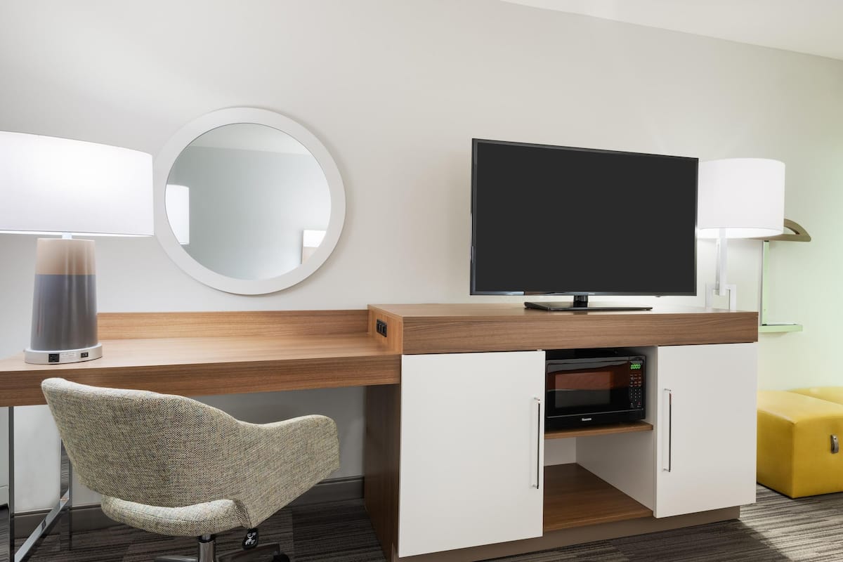 Una habitación de hotel moderna con un escritorio de madera, un televisor de pantalla plana, un espejo redondo, una lámpara, un microondas y un otoman amarillo.
