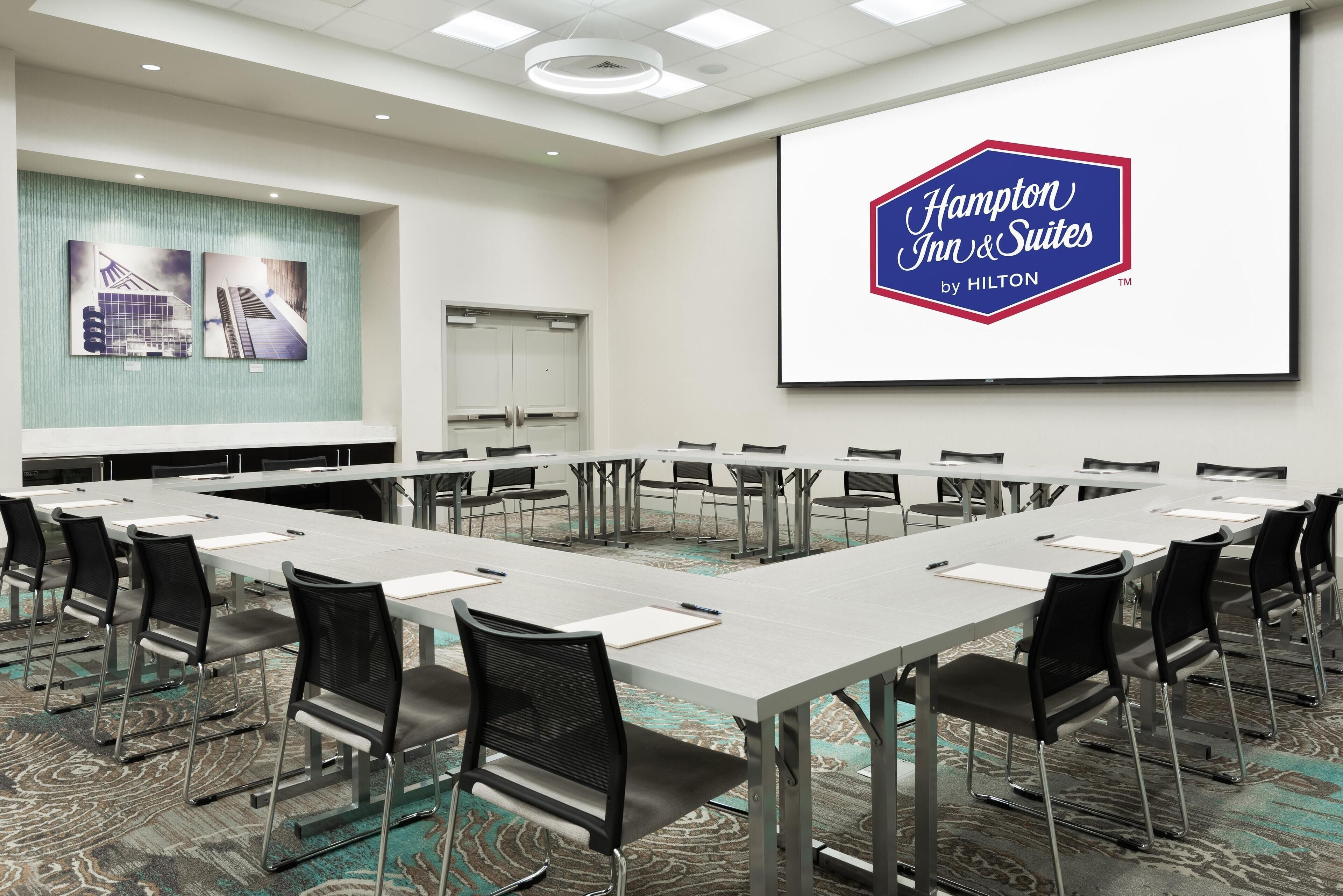 A conference room with a long table and chairs, a large screen displaying the Hampton Inn & Suites logo, and a carpeted floor.