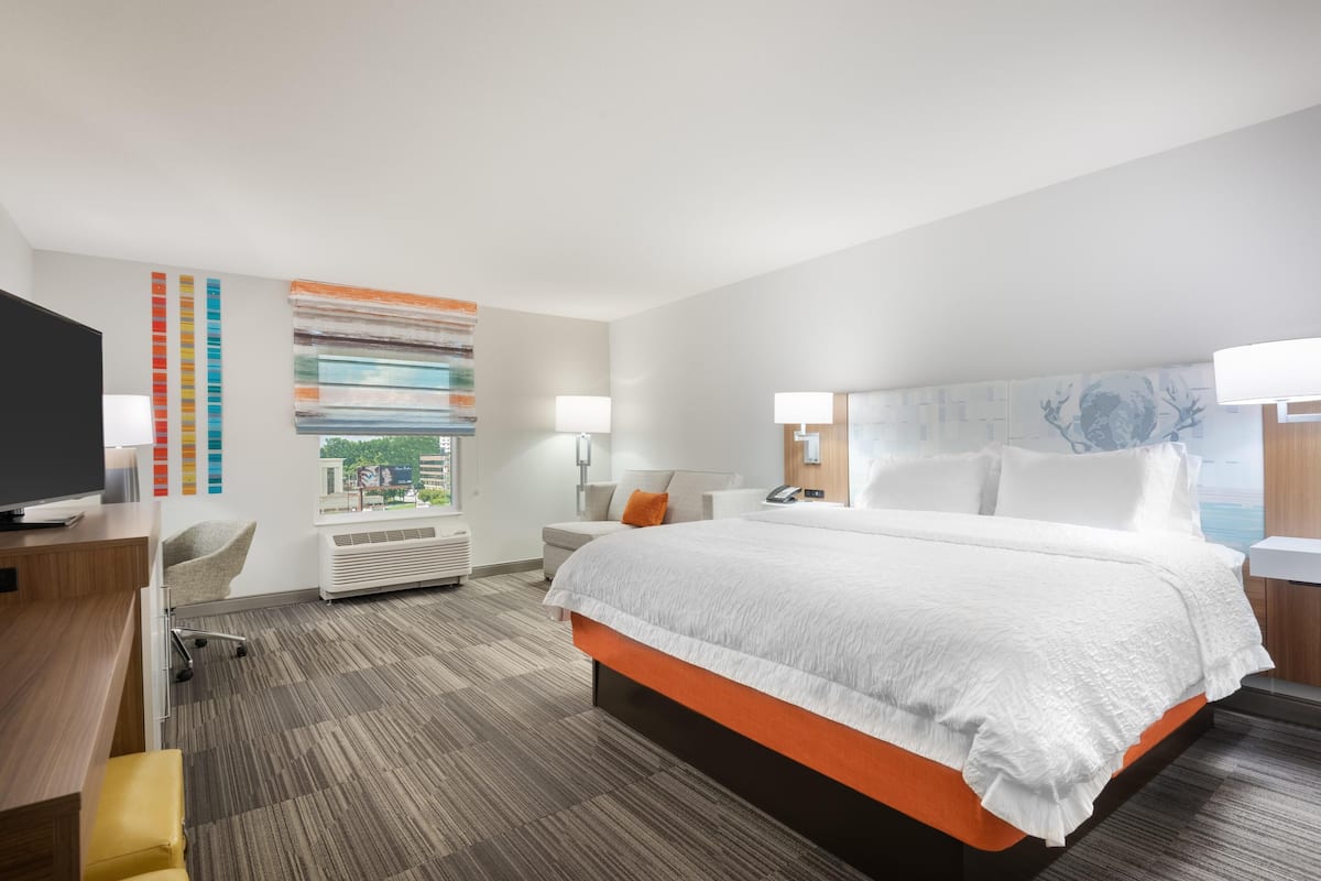 Habitación de hotel con una cama grande, un escritorio con computadora, una silla, un televisor y una ventana con vista a la ciudad.