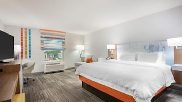 Habitación de hotel con una cama grande, un escritorio con computadora, una silla, un televisor y una ventana con vista a la ciudad.
