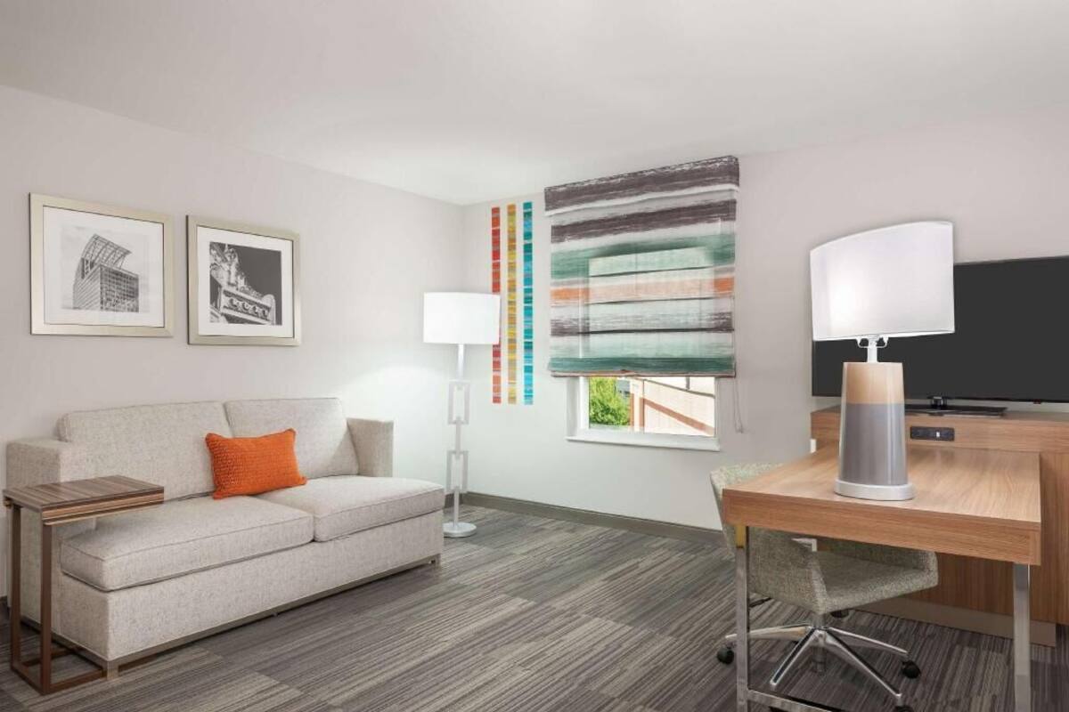 Una habitación de hotel moderna con sofá, escritorio, lámpara y un cuadro abstracto grande.