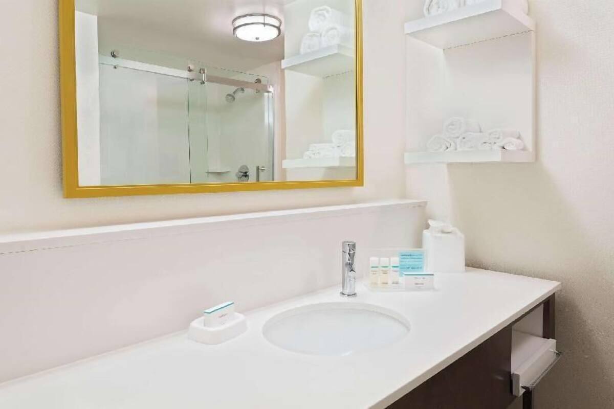 Un baño con un espejo grande, un lavamanos y repisas con artículos de aseo.
