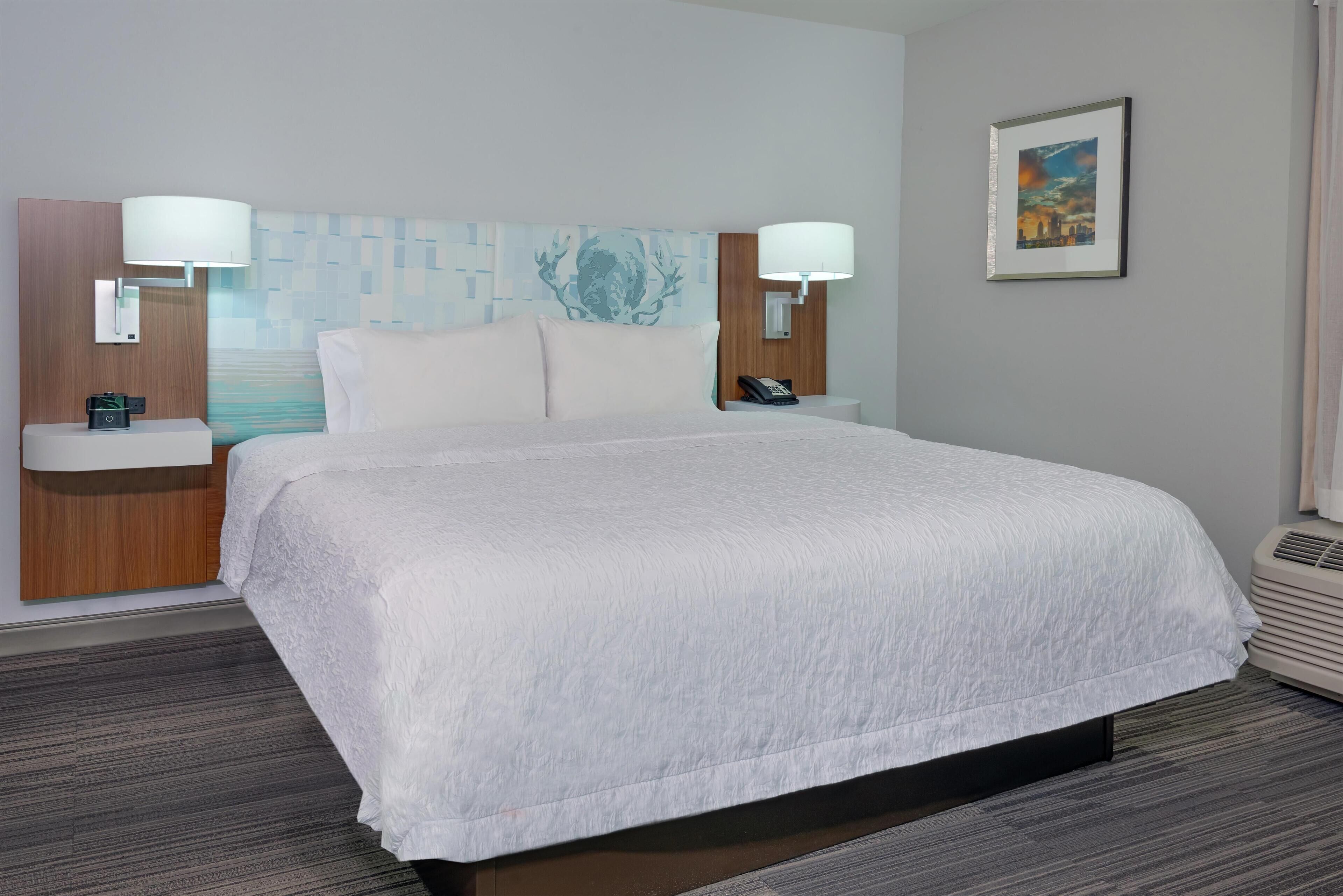 A hotel room with a large bed, two bedside tables with lamps, a wall-mounted phone, a framed picture, and a carpeted floor.