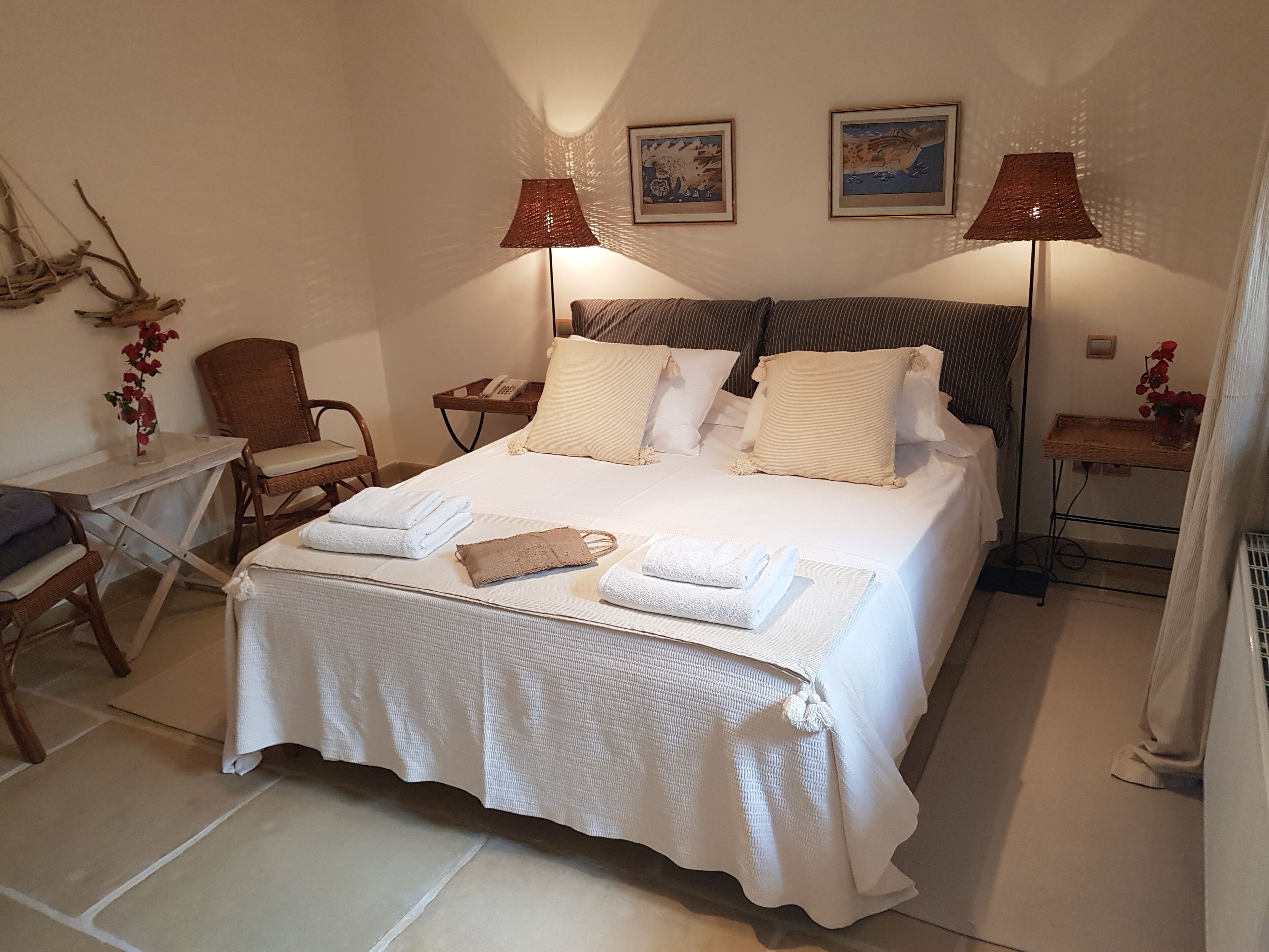 A neatly made bed with white linens and a brown headboard, a small table with a phone and a vase of flowers, and two wall-mounted lamps.