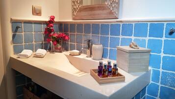Une salle de bain avec un lavabo blanc, des carreaux de mosaïque bleus, un vase rempli de fleurs rouges et un plateau en bois contenant des bouteilles.
