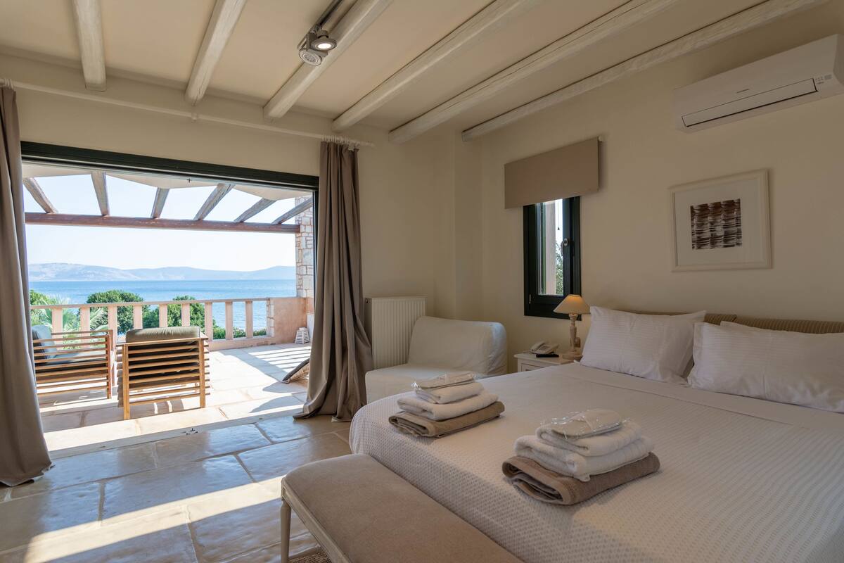 A bedroom with a large bed, a chair, a nightstand, and a view of the sea.