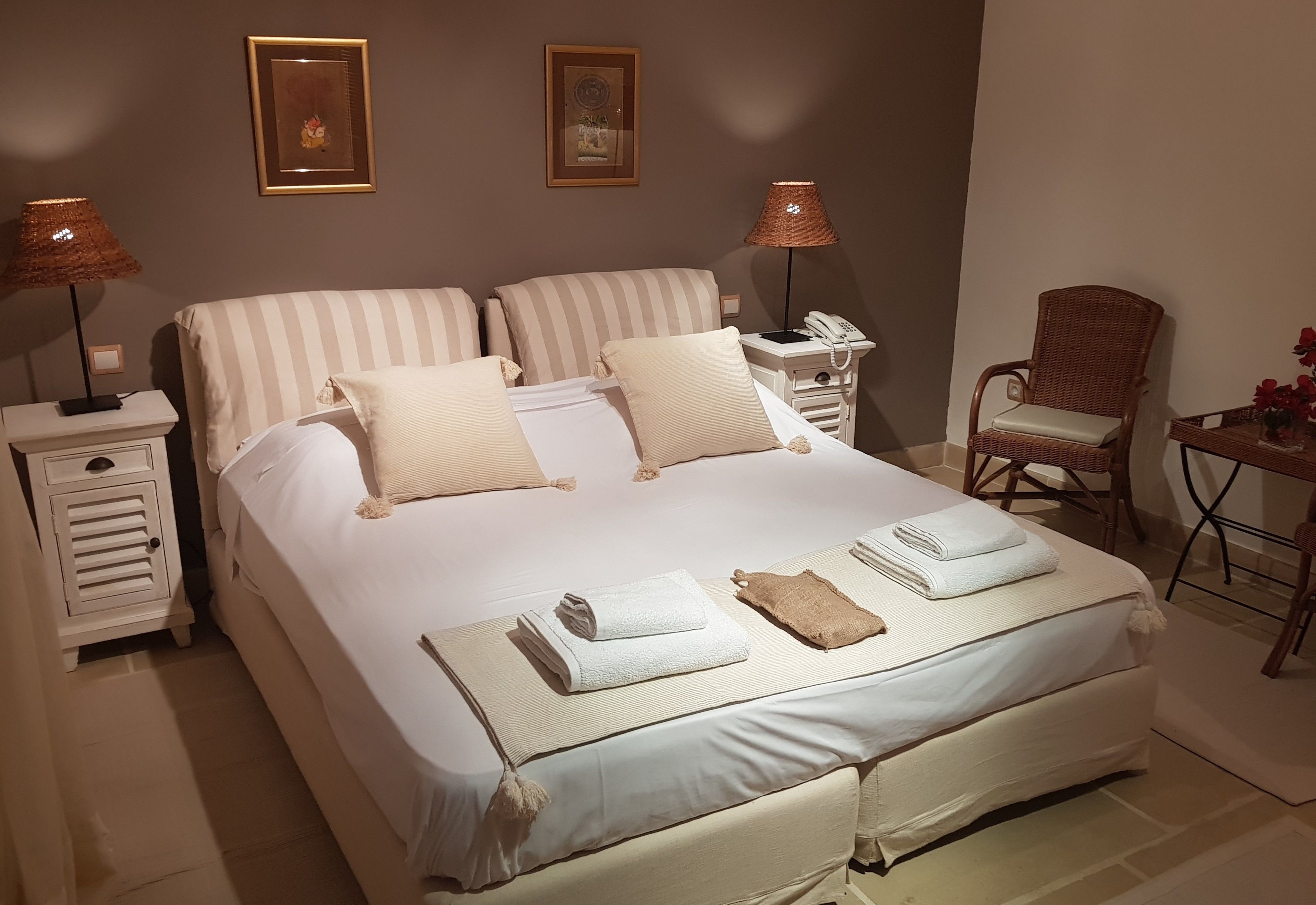 A double bed with white and beige bedding, two bedside tables with lamps, a wicker chair, and a small table with a vase.