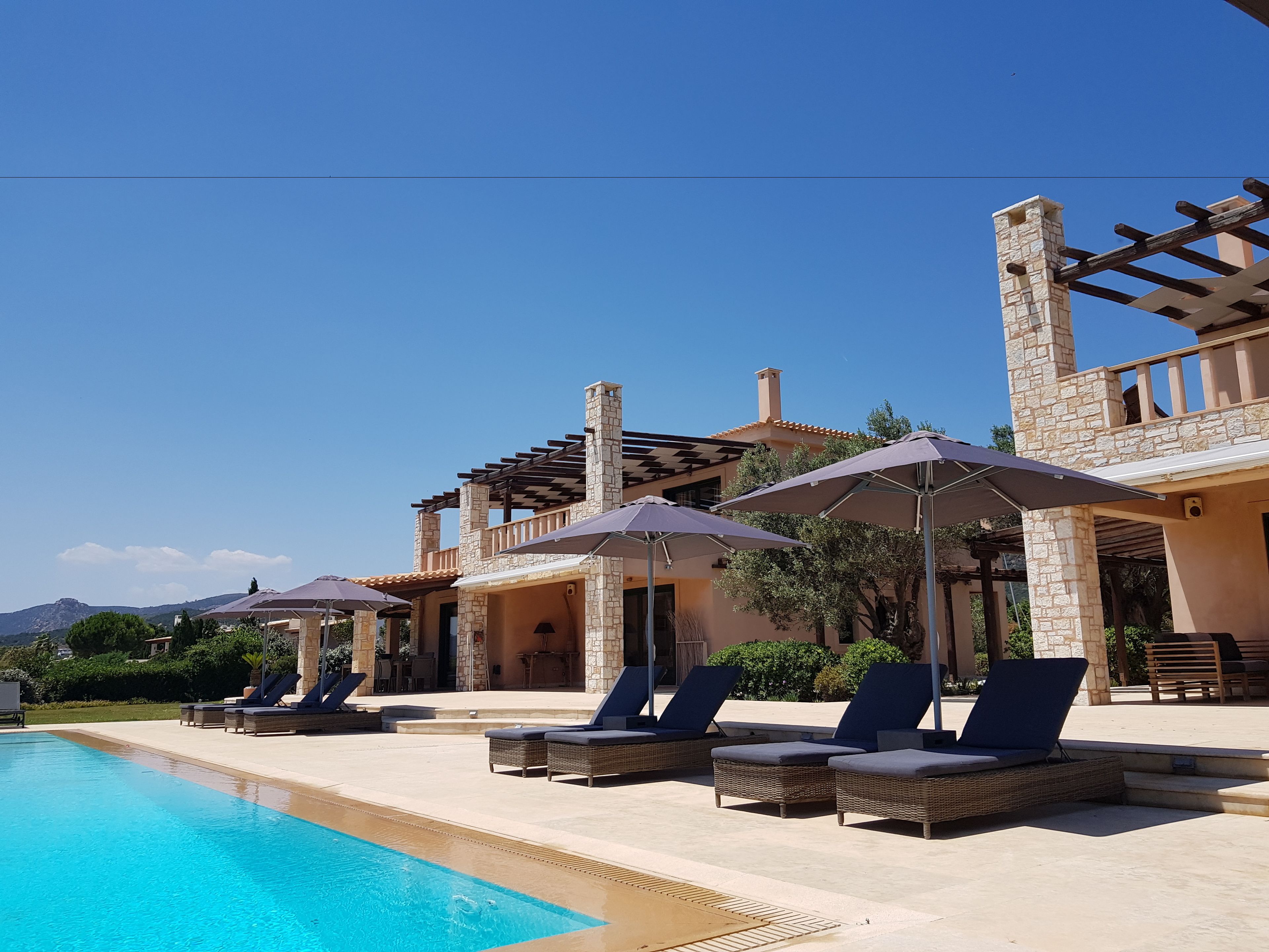 A poolside area with lounge chairs and umbrellas, a stone building with a balcony, and a clear blue sky.