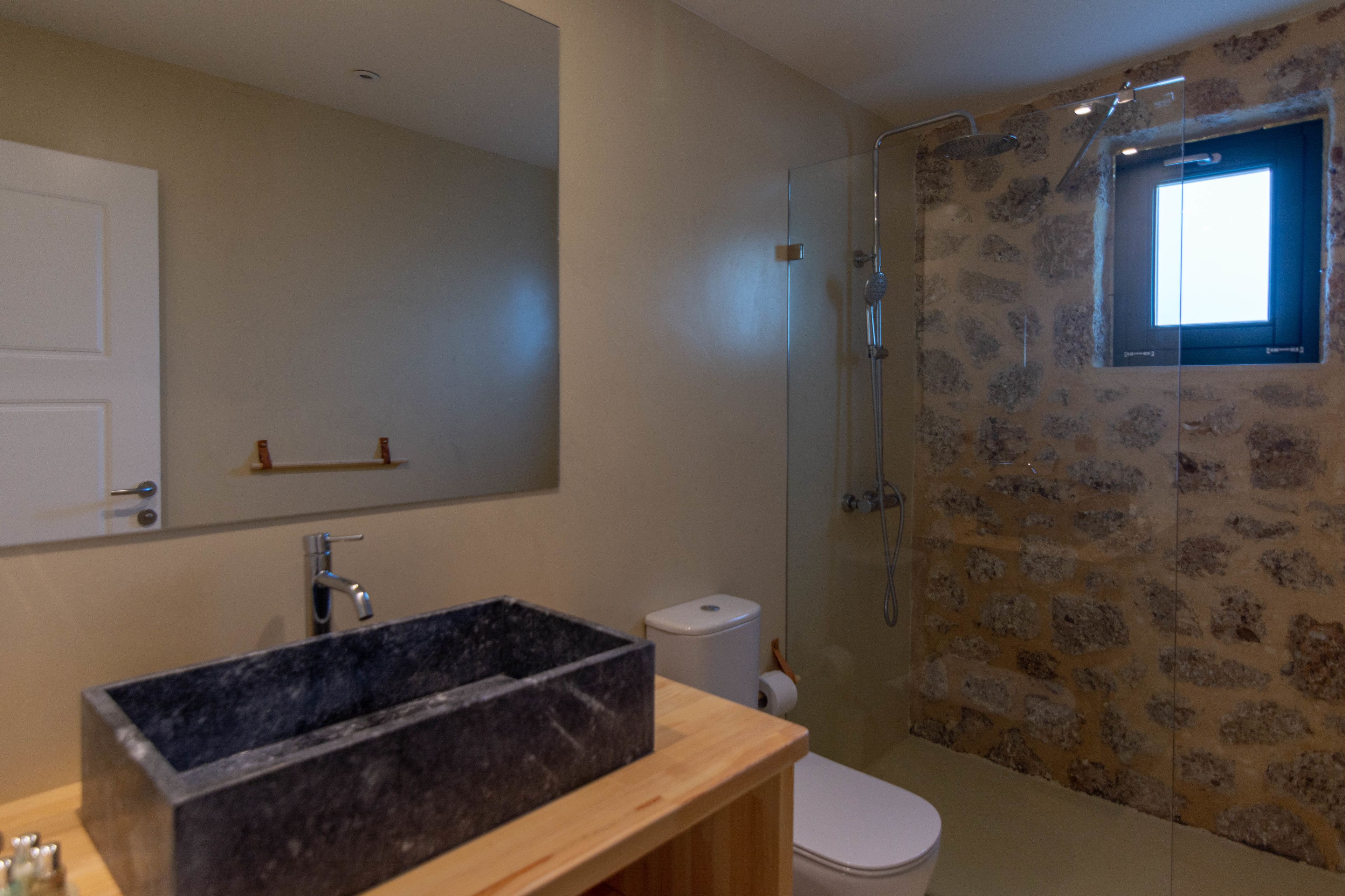 A bathroom with a large mirror, a black sink, a toilet, and a shower with a stone wall.