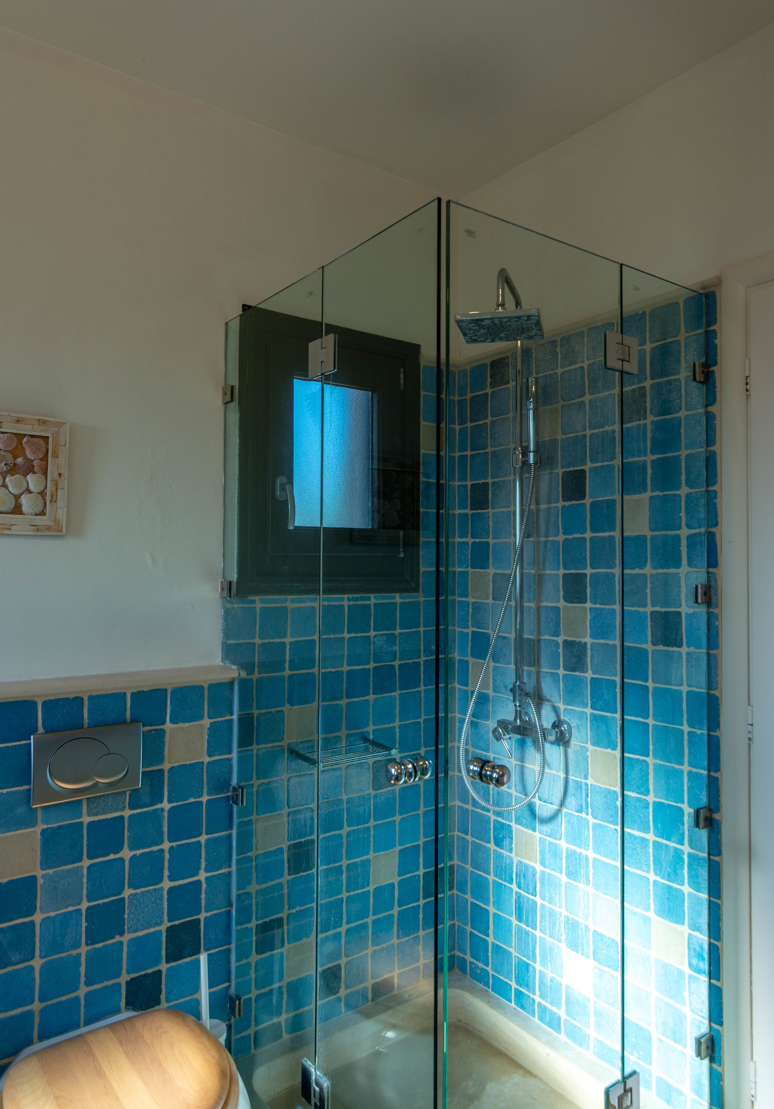A glass-enclosed shower with blue tiles, a wooden toilet, and a small shelf with a decorative item.