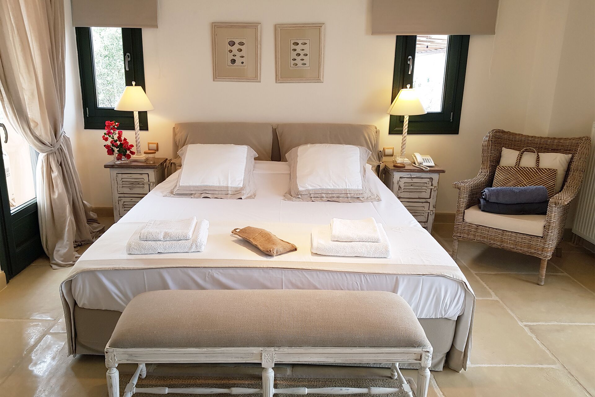 A well-lit bedroom with a large bed, two bedside tables, a wicker chair, and a bench.