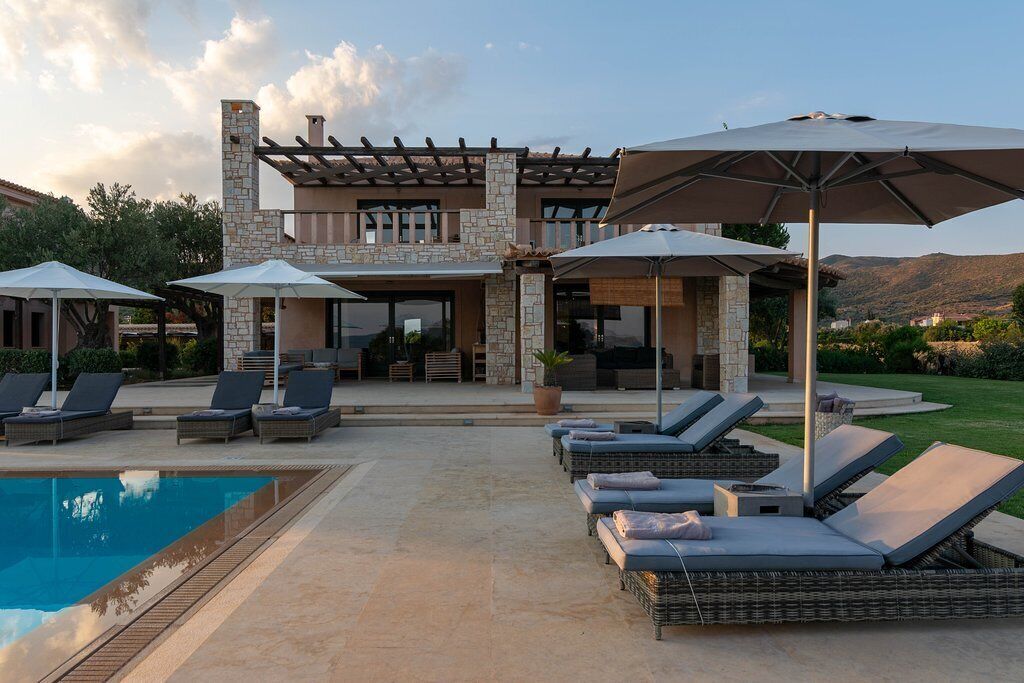 Zona junto a la piscina con sillones de descanso y sombrillas, un edificio de piedra con pérgola y vistas a las montañas.
